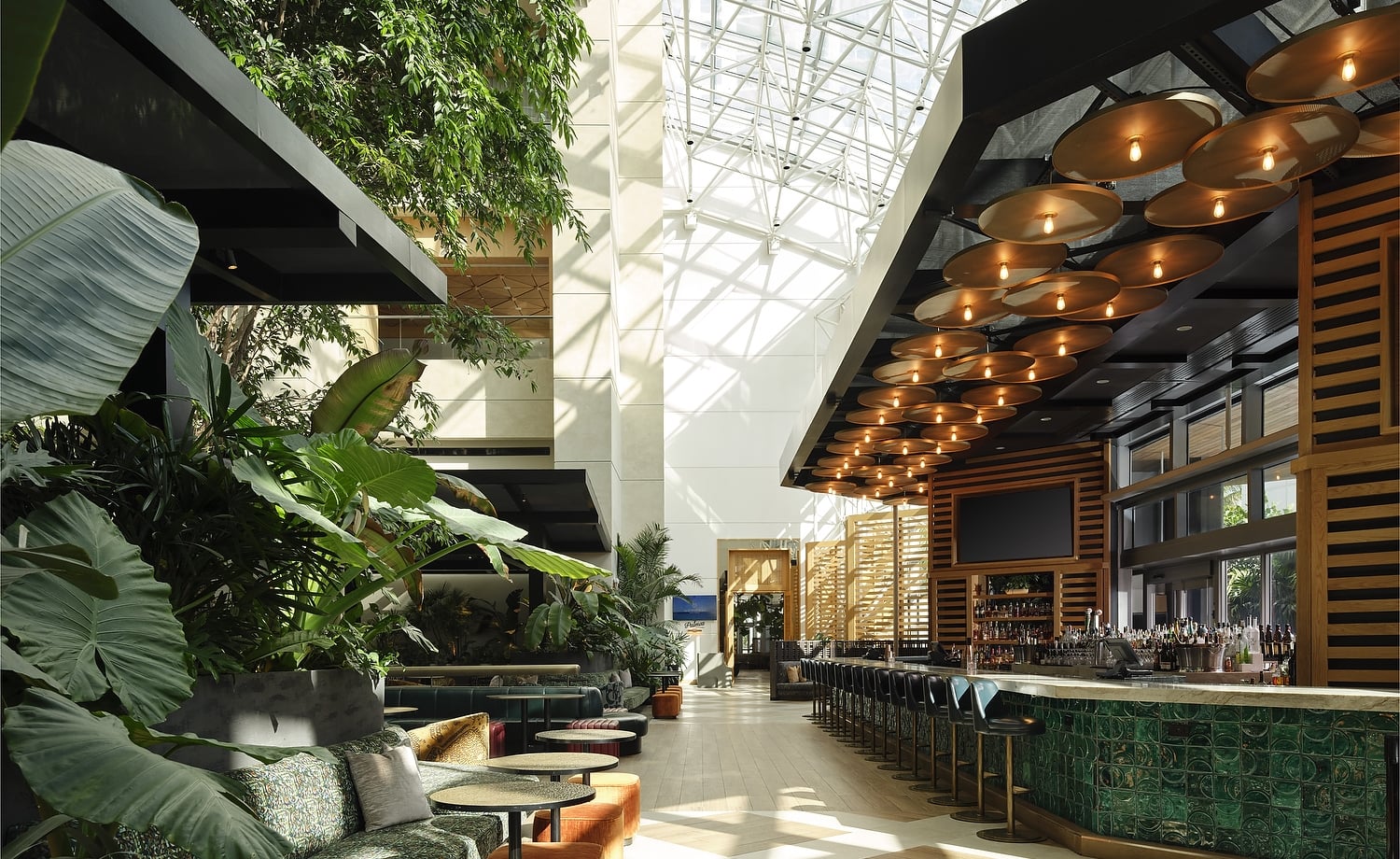 A spacious, modern hotel lounge features a long, green tiled bar and abundant tropical plants providing a lush seating area. Natural light streams from a high glass atrium ceiling. Text: EXIT, Palm Court.