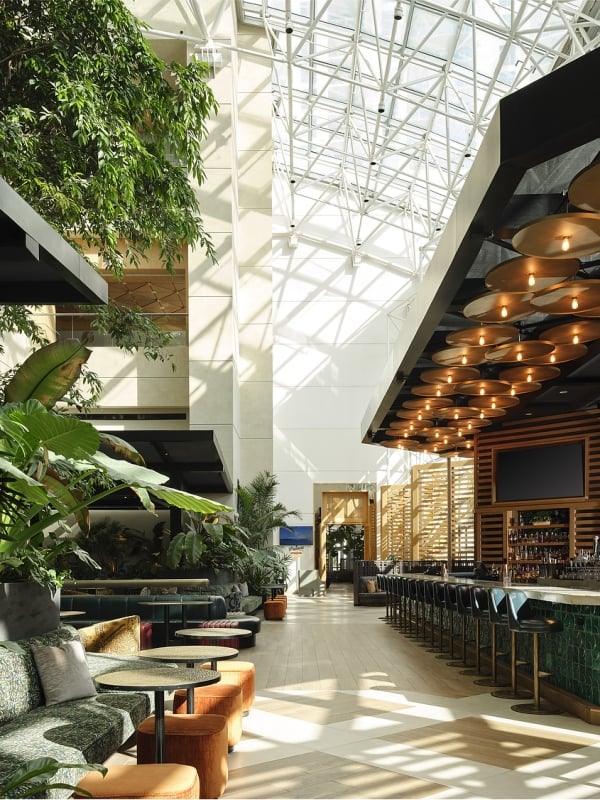 A spacious, modern hotel lounge features a long, green tiled bar and abundant tropical plants providing a lush seating area. Natural light streams from a high glass atrium ceiling. Text: EXIT, Palm Court.