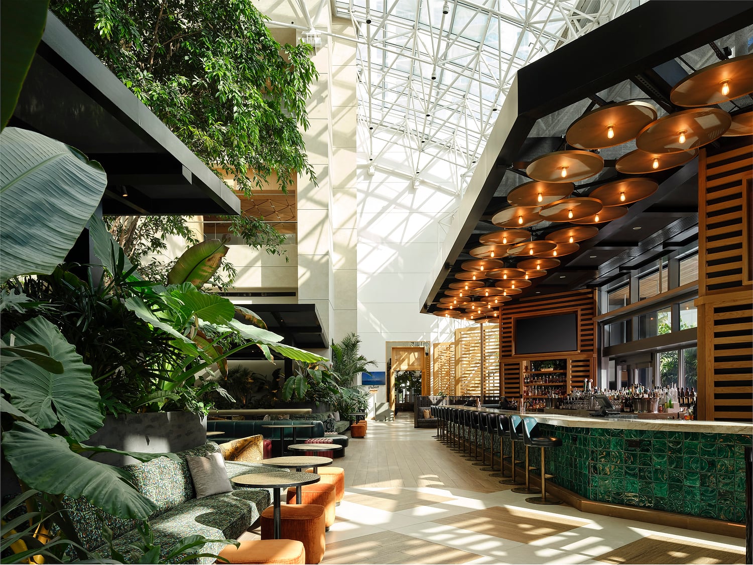 A spacious, modern hotel lounge features a long, green tiled bar and abundant tropical plants providing a lush seating area. Natural light streams from a high glass atrium ceiling. Text: EXIT, Palm Court.