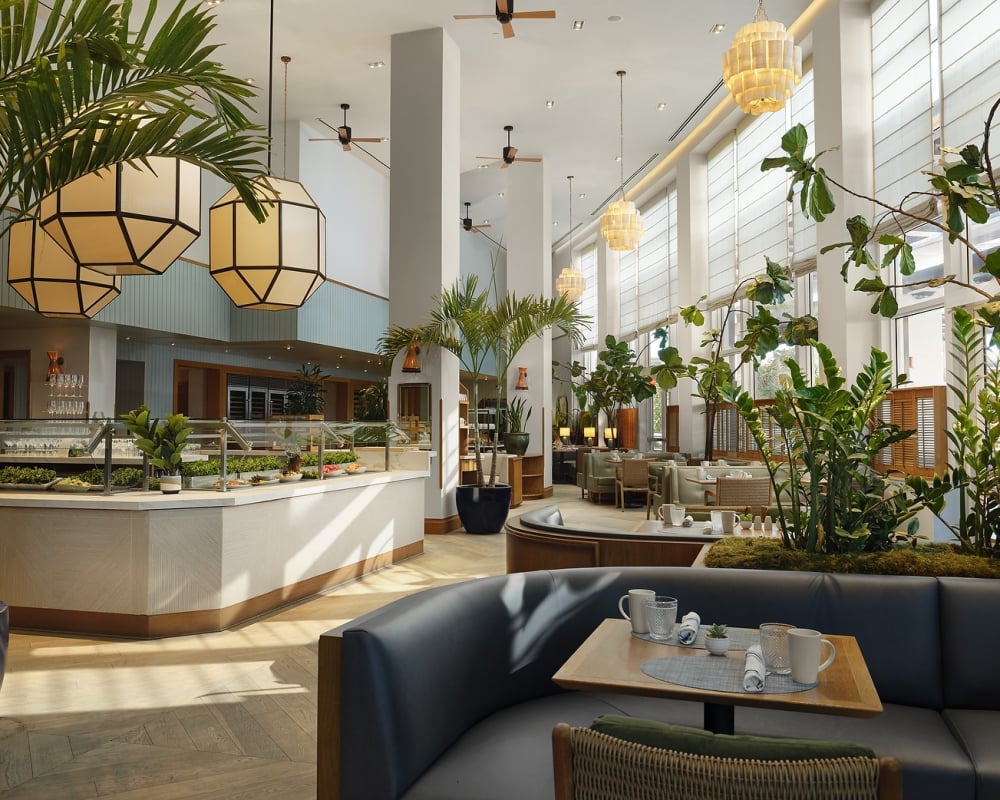 A spacious, modern restaurant features a buffet counter and set tables amidst abundant indoor plants, high ceilings, and large windows with blinds.
