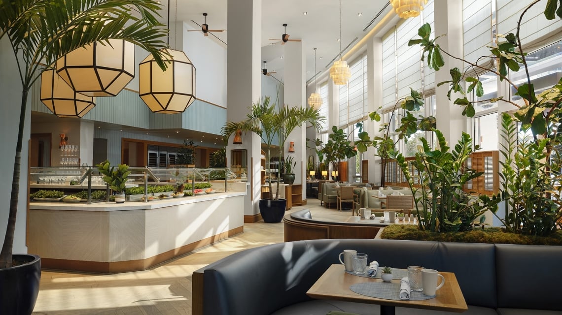 A spacious, modern restaurant features a buffet counter and set tables amidst abundant indoor plants, high ceilings, and large windows with blinds.