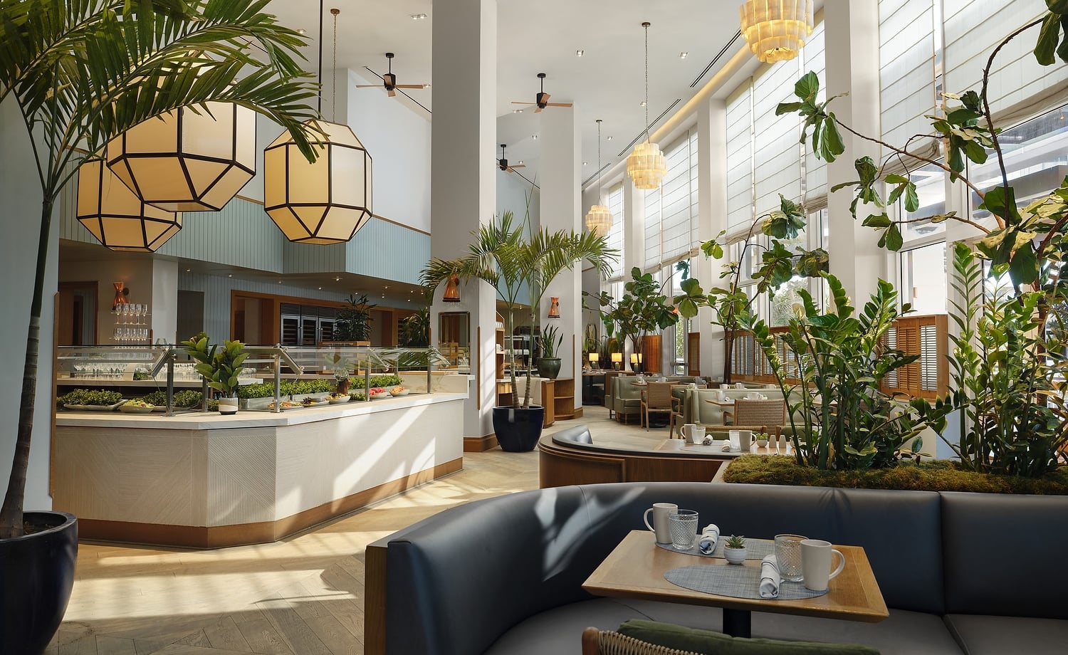 A spacious, modern restaurant features a buffet counter and set tables amidst abundant indoor plants, high ceilings, and large windows with blinds.