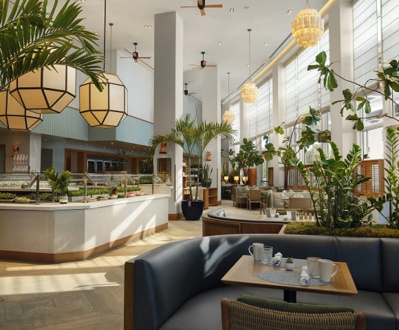A spacious, modern restaurant features a buffet counter and set tables amidst abundant indoor plants, high ceilings, and large windows with blinds.