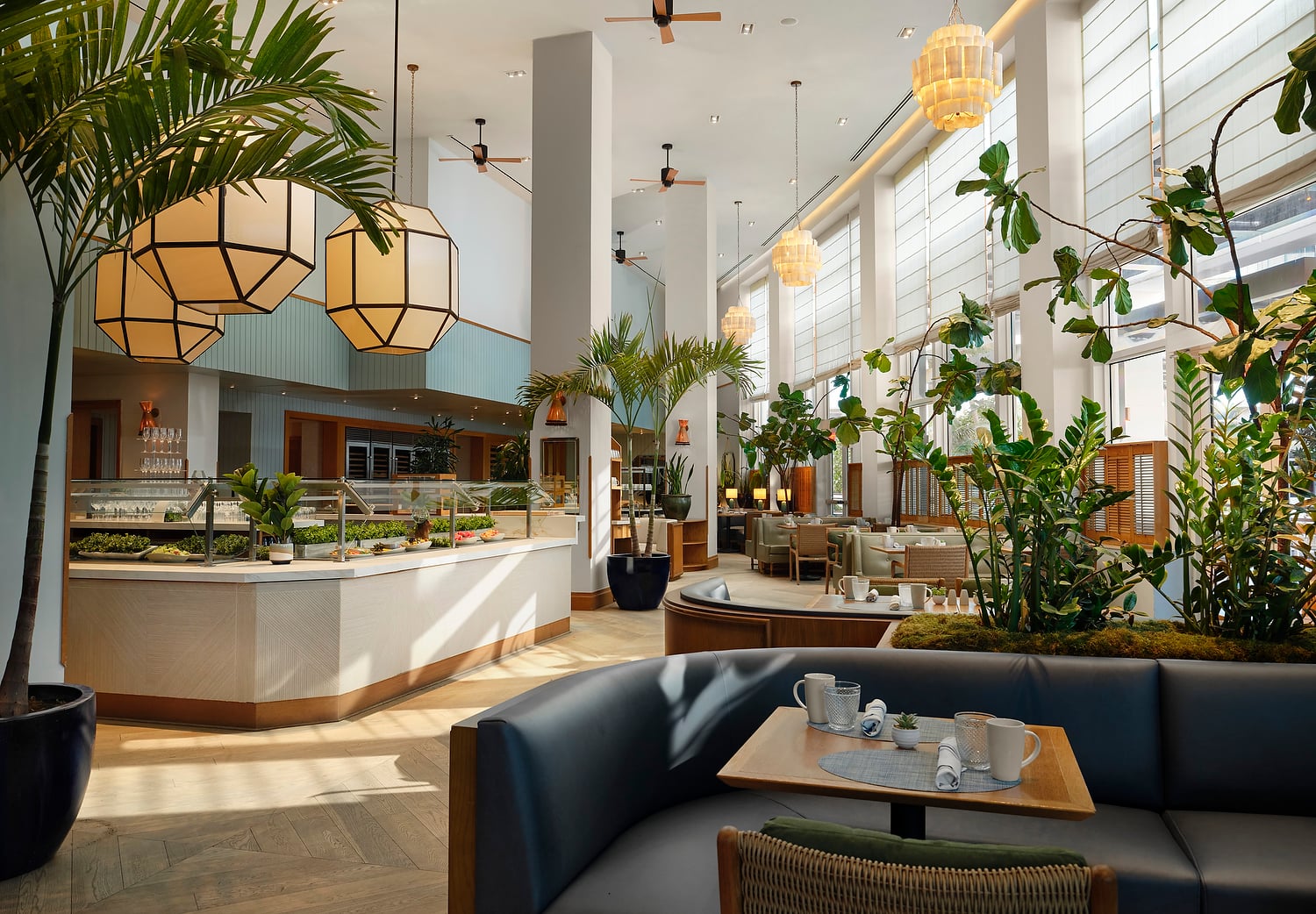 A spacious, modern restaurant features a buffet counter and set tables amidst abundant indoor plants, high ceilings, and large windows with blinds.