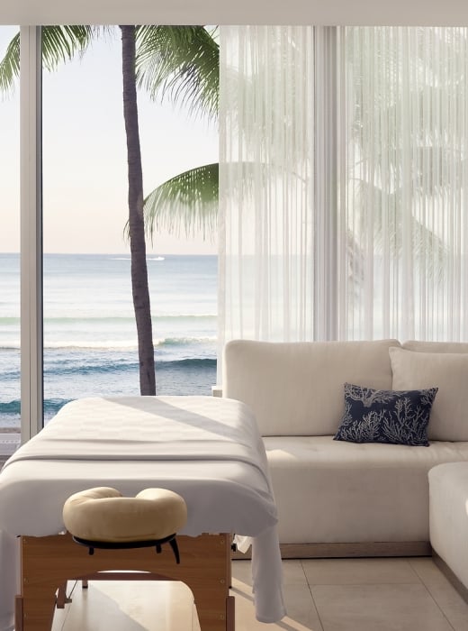 Two massage tables are set up in a bright room with a large window overlooking a calm ocean, beach, and palm tree, beside a light-colored sectional sofa.