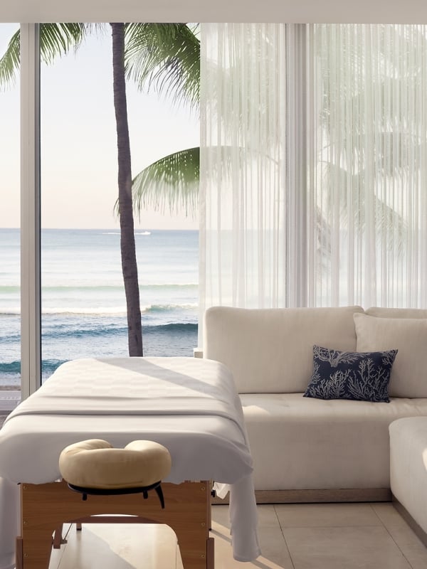 Two massage tables are set up in a bright room with a large window overlooking a calm ocean, beach, and palm tree, beside a light-colored sectional sofa.