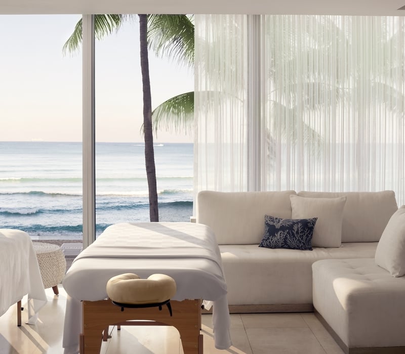 Two massage tables are set up in a bright room with a large window overlooking a calm ocean, beach, and palm tree, beside a light-colored sectional sofa.