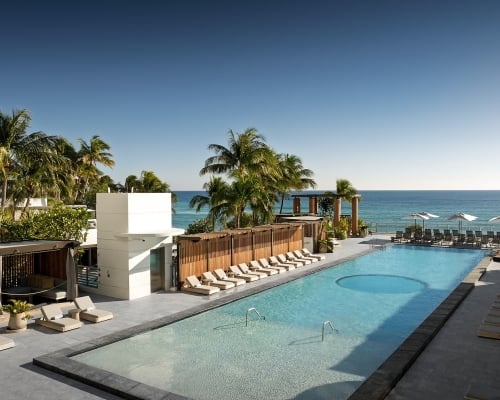 A large resort pool with lounge chairs and cabanas sits beside palm trees, overlooking the blue ocean under a clear sky.