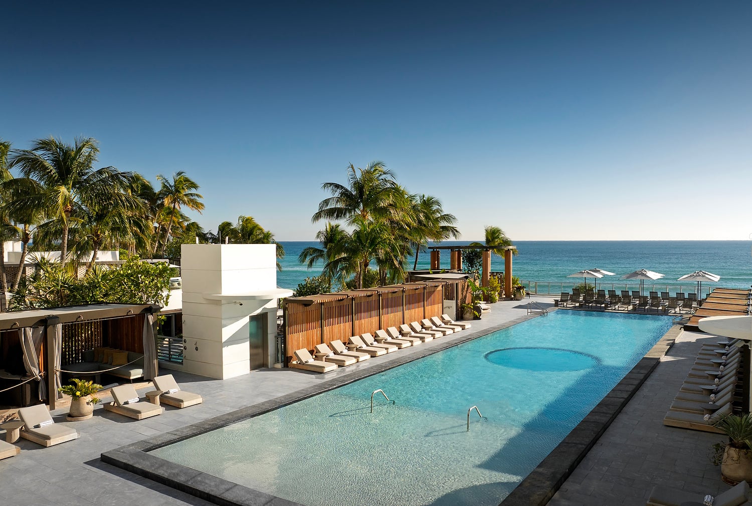 A large resort pool with lounge chairs and cabanas sits beside palm trees, overlooking the blue ocean under a clear sky.