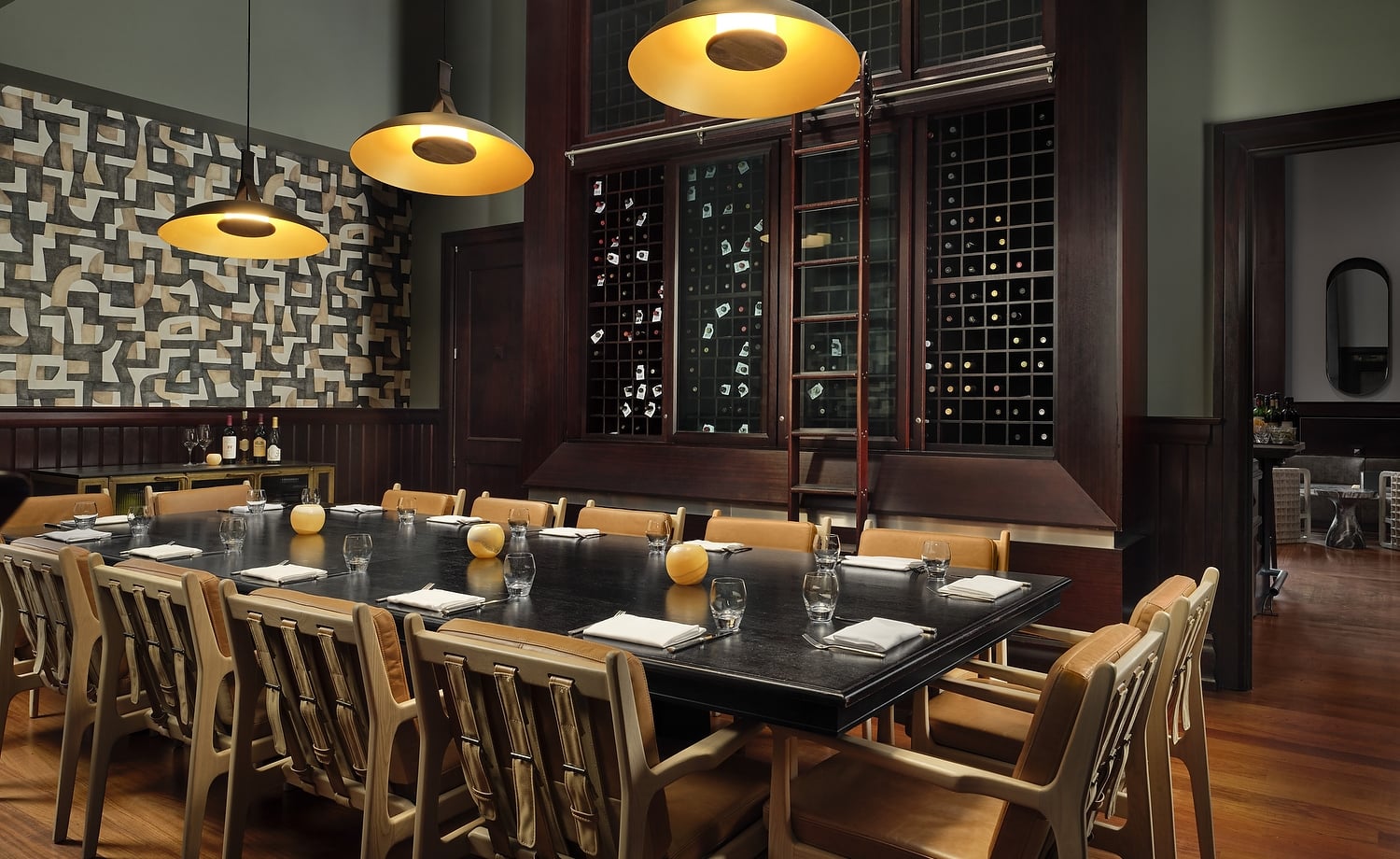 A long black dining table is set with glasses and napkins, surrounded by tan leather chairs. Dark wood wine racks with a sliding ladder dominate the background, illuminated by three golden pendant lights.