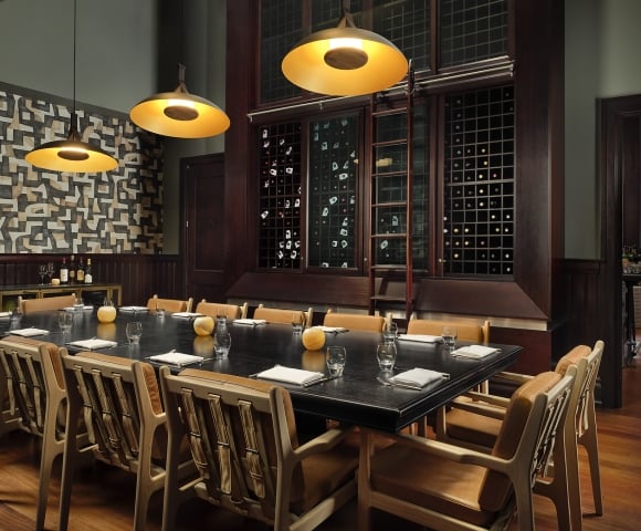 A long black dining table is set with glasses and napkins, surrounded by tan leather chairs. Dark wood wine racks with a sliding ladder dominate the background, illuminated by three golden pendant lights.