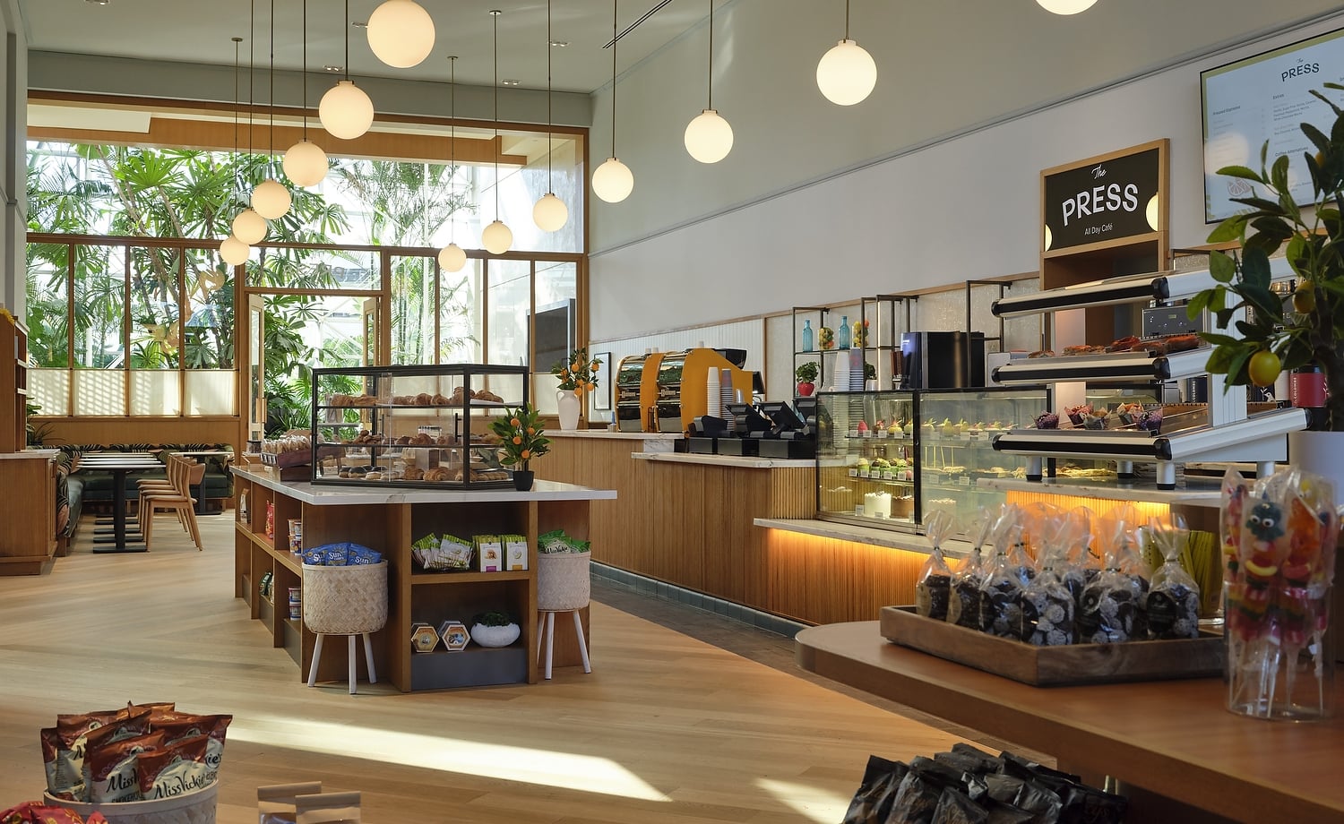 Bright "The PRESS All Day Café" features counters with pastries and packaged goods. Large windows reveal lush greenery. Menu lists: "PRESS," "Espresso," "Coffee alternatives," "Cold press."