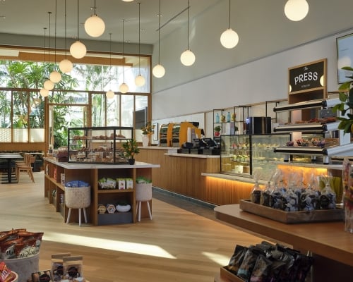 Bright "The PRESS All Day Café" features counters with pastries and packaged goods. Large windows reveal lush greenery. Menu lists: "PRESS," "Espresso," "Coffee alternatives," "Cold press."
