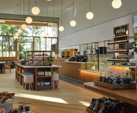 Bright "The PRESS All Day Café" features counters with pastries and packaged goods. Large windows reveal lush greenery. Menu lists: "PRESS," "Espresso," "Coffee alternatives," "Cold press."