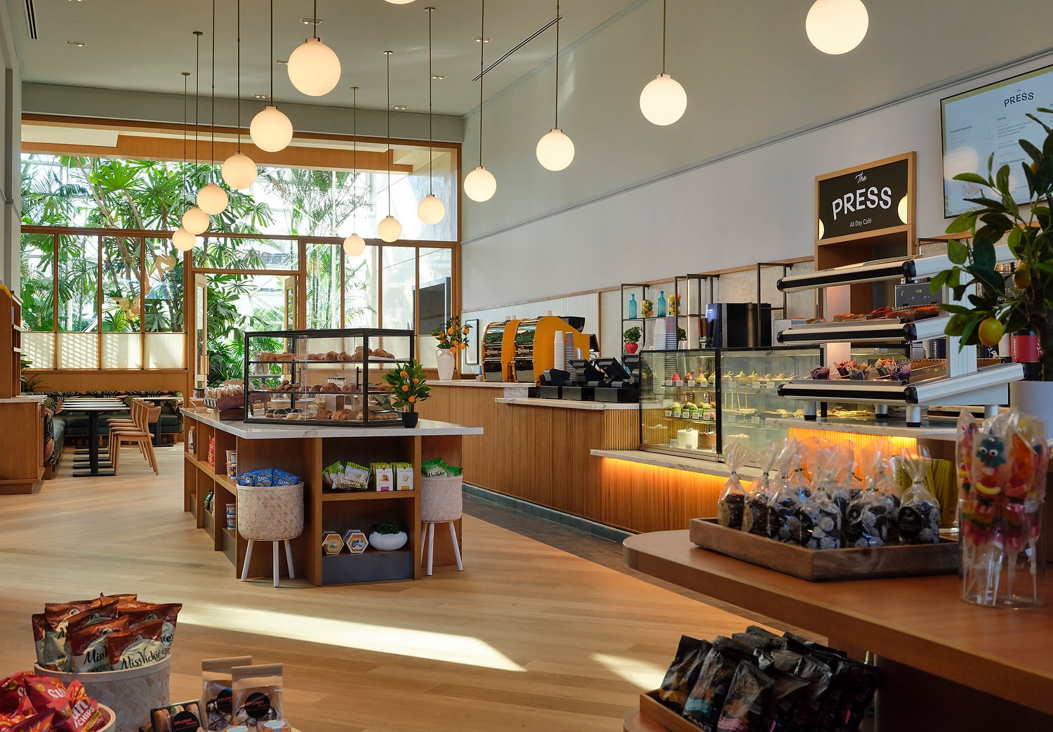 Bright "The PRESS All Day Café" features counters with pastries and packaged goods. Large windows reveal lush greenery. Menu lists: "PRESS," "Espresso," "Coffee alternatives," "Cold press."