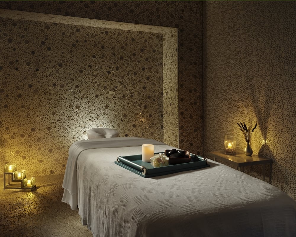 A white-blanketed massage table holds a tray with a lit candle, flower, and stones. It is in a dimly lit spa room with walls and floor of small round wooden discs.