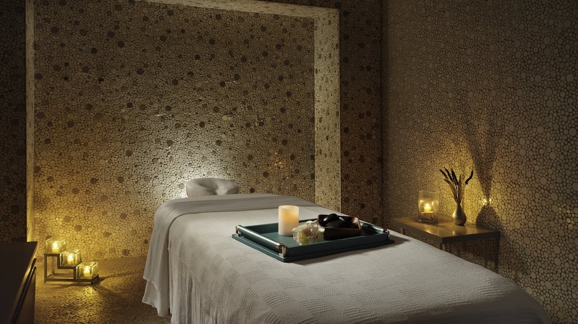 A white-blanketed massage table holds a tray with a lit candle, flower, and stones. It is in a dimly lit spa room with walls and floor of small round wooden discs.