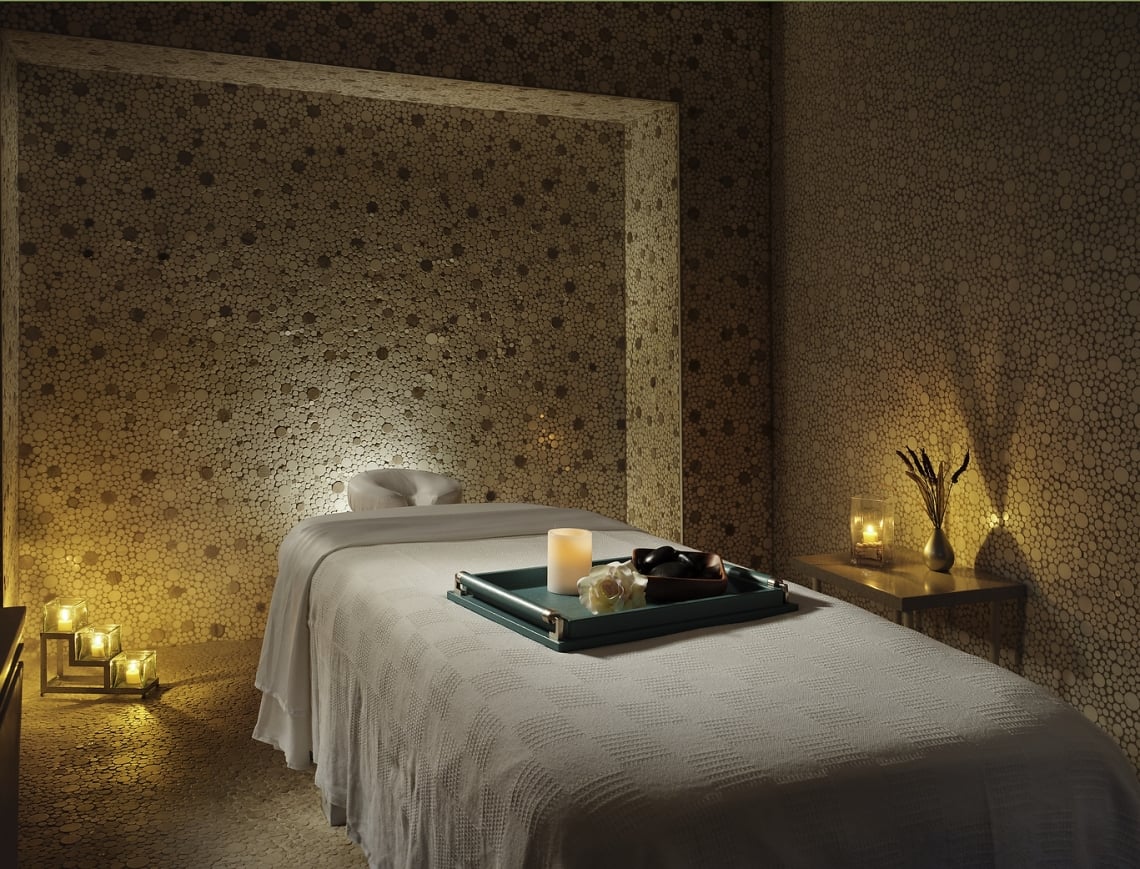 A white-blanketed massage table holds a tray with a lit candle, flower, and stones. It is in a dimly lit spa room with walls and floor of small round wooden discs.