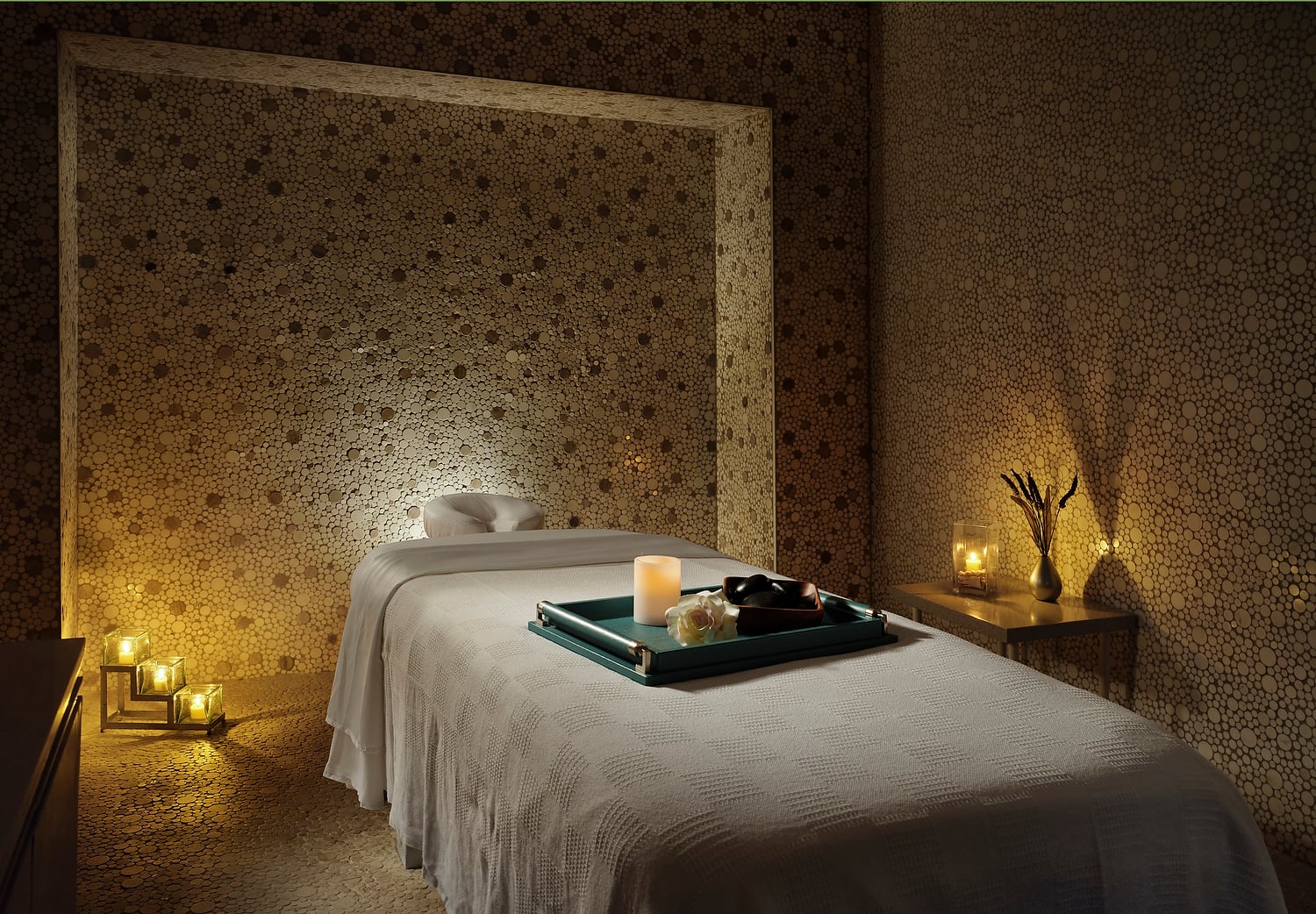 A white-blanketed massage table holds a tray with a lit candle, flower, and stones. It is in a dimly lit spa room with walls and floor of small round wooden discs.