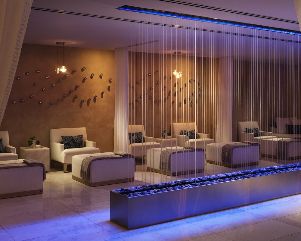 A modern spa relaxation room features multiple inviting white armchairs and ottomans arranged around a blue-lit pebble water feature, with decorative wall art and warm overhead lights.