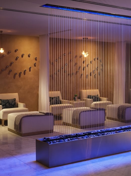 A modern spa relaxation room features multiple inviting white armchairs and ottomans arranged around a blue-lit pebble water feature, with decorative wall art and warm overhead lights.