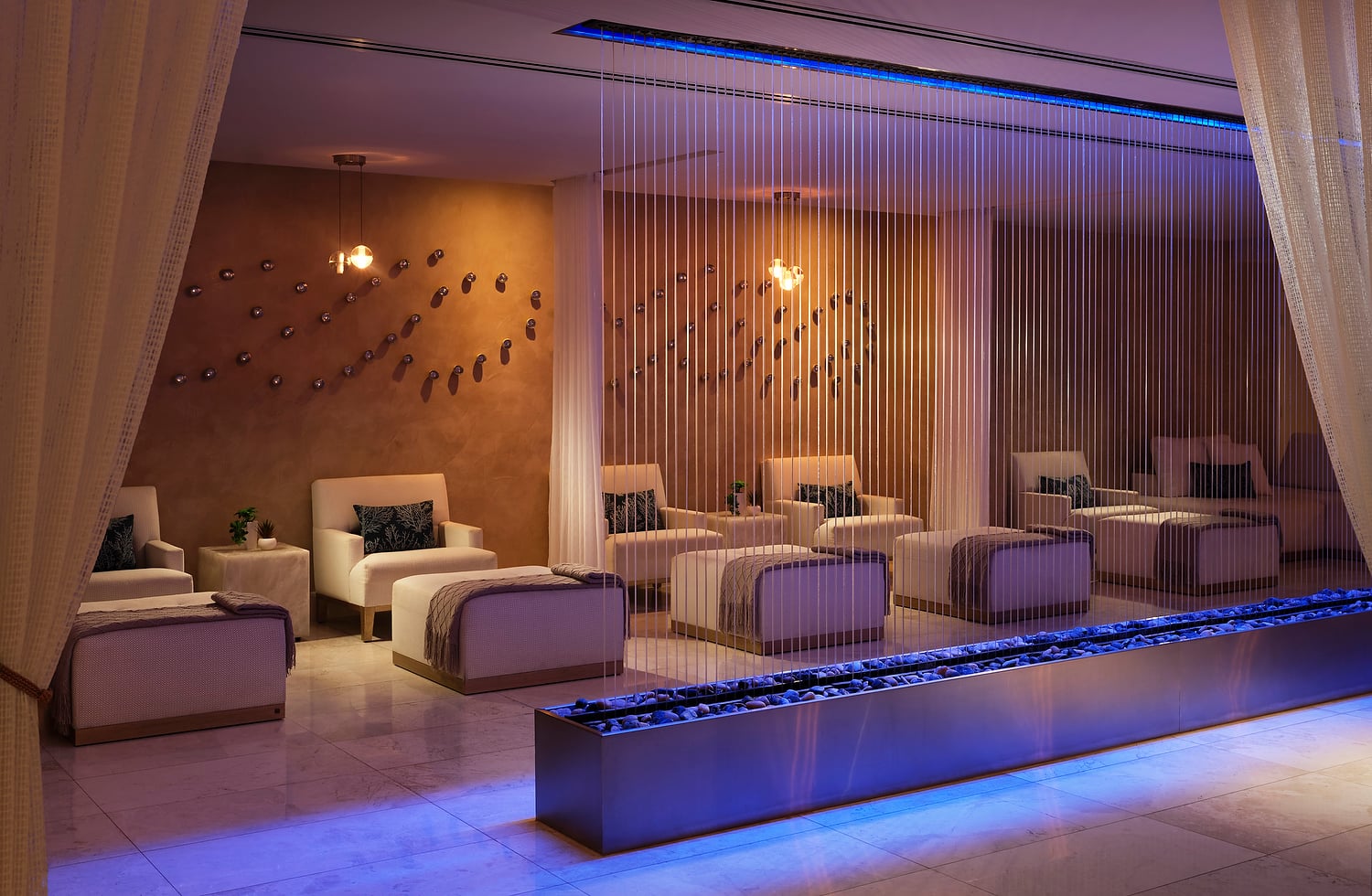 A modern spa relaxation room features multiple inviting white armchairs and ottomans arranged around a blue-lit pebble water feature, with decorative wall art and warm overhead lights.