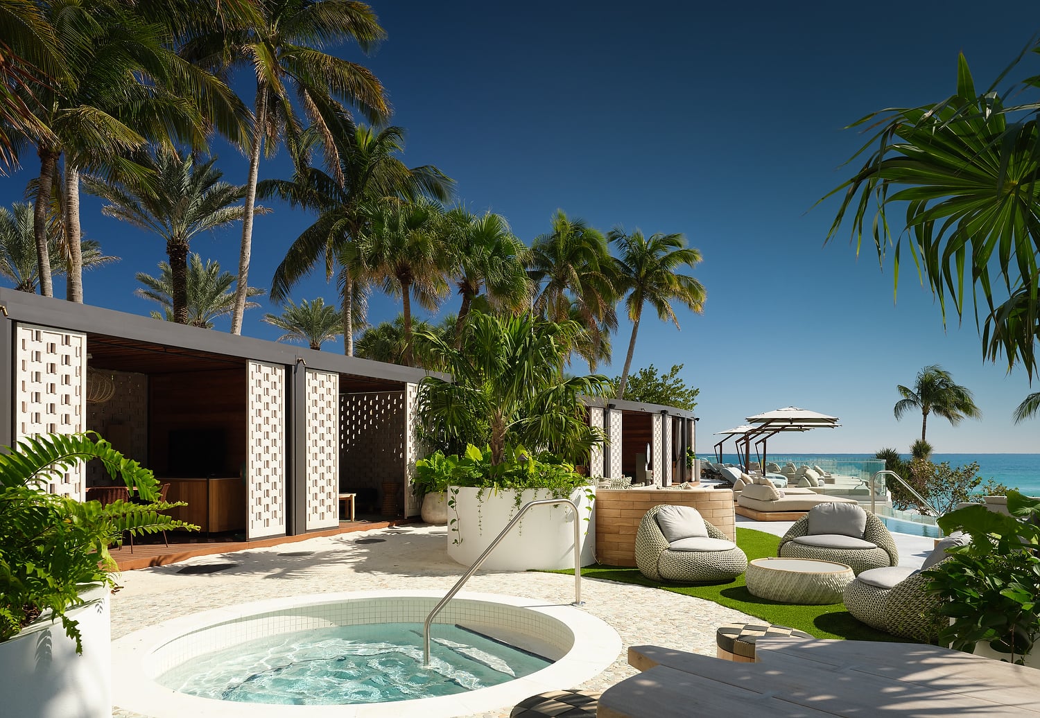 A resort-style outdoor lounge area features a bubbling hot tub, modern cabanas, and wicker seating, set under palm trees with a clear blue sky overlooking a turquoise ocean.