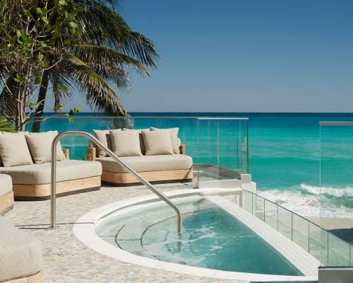 A curved hot tub and beige sofas sit on a pebble patio, overlooking a vibrant turquoise ocean and sandy beach. Palm trees and glass railings frame the clear blue sky view.