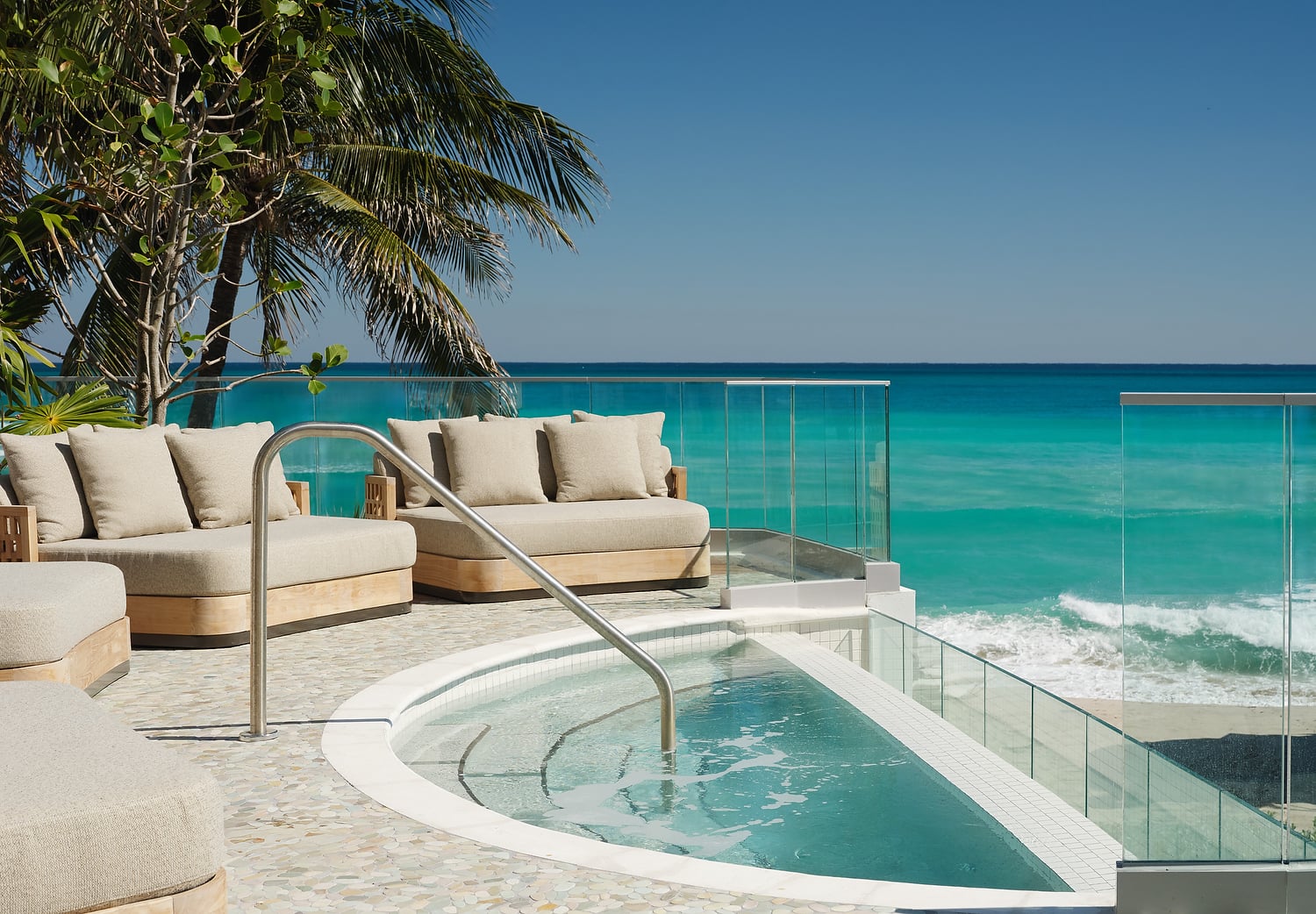 A curved hot tub and beige sofas sit on a pebble patio, overlooking a vibrant turquoise ocean and sandy beach. Palm trees and glass railings frame the clear blue sky view.