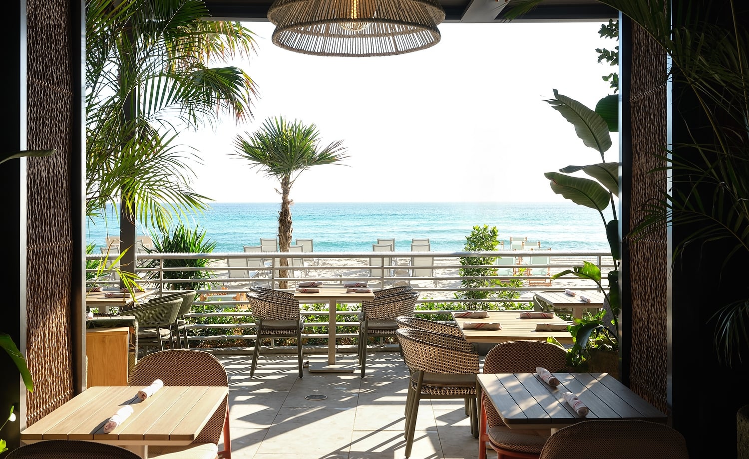 An outdoor restaurant patio with set tables overlooks a bright beach and blue ocean, framed by lush palm trees. A large woven light fixture hangs overhead.