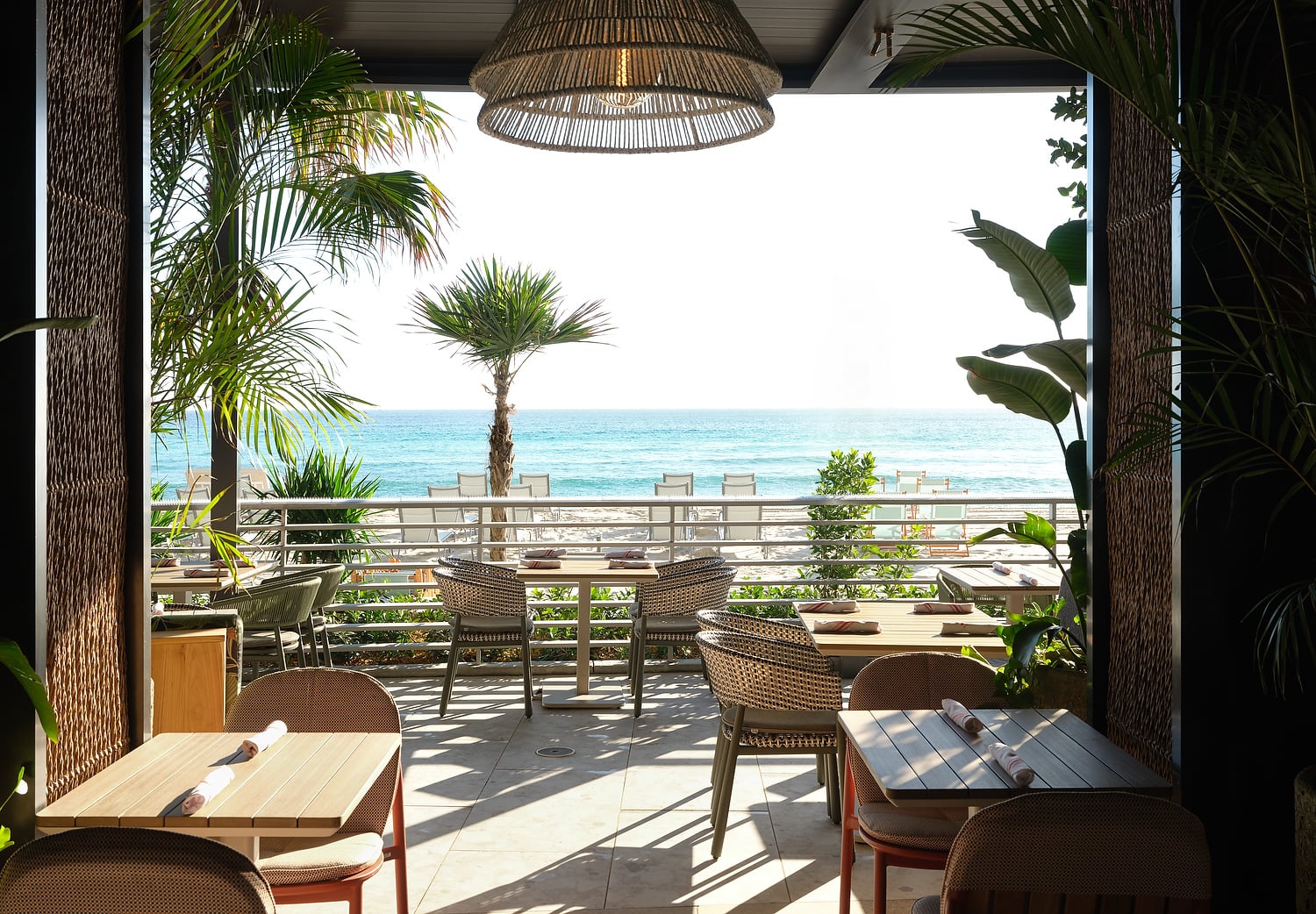 An outdoor restaurant patio with set tables overlooks a bright beach and blue ocean, framed by lush palm trees. A large woven light fixture hangs overhead.