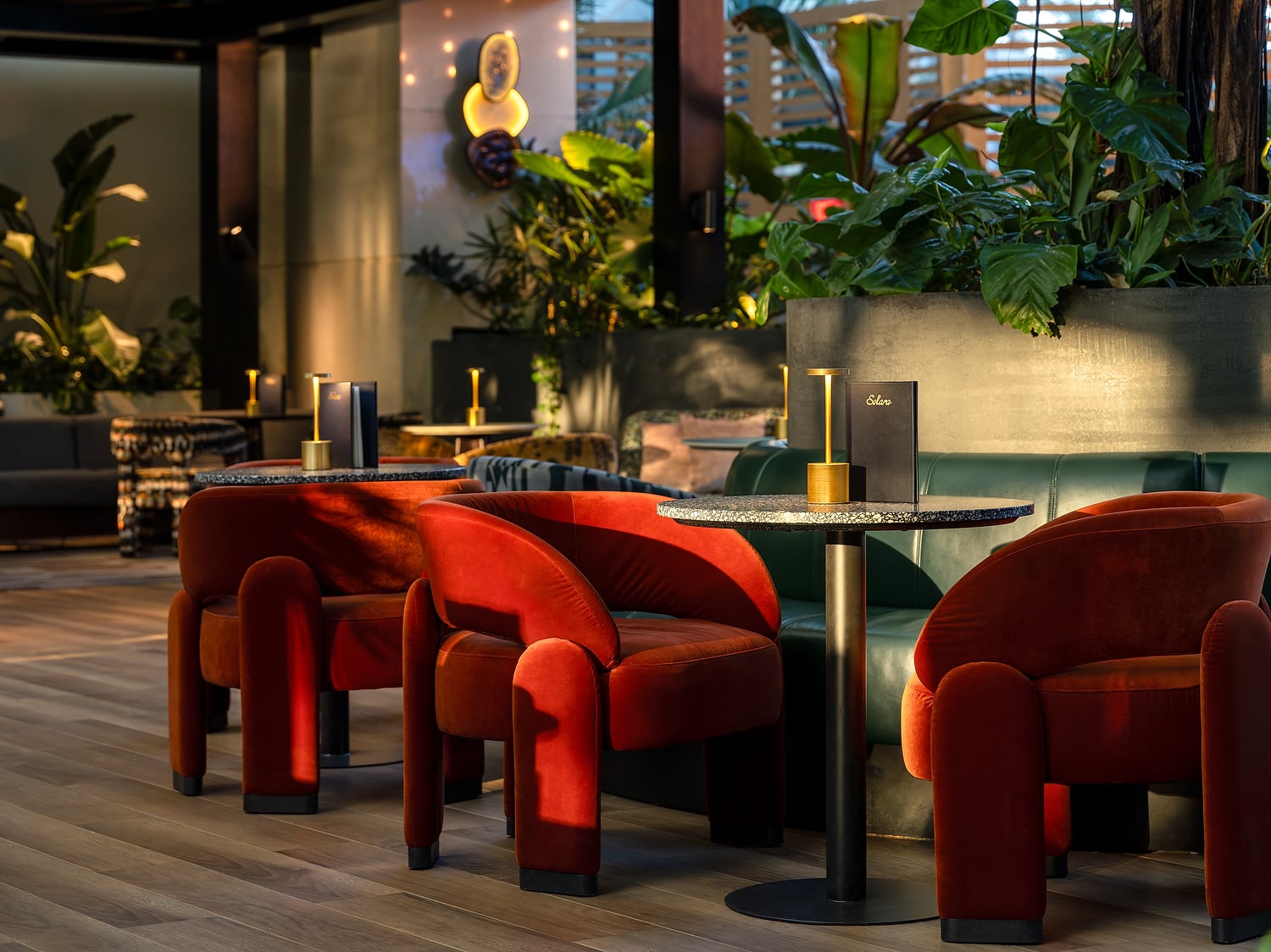 Plush red velvet armchairs and small tables offer seating in a modern, plant-filled lounge with warm accent lighting. A menu on a table reads "Solara".