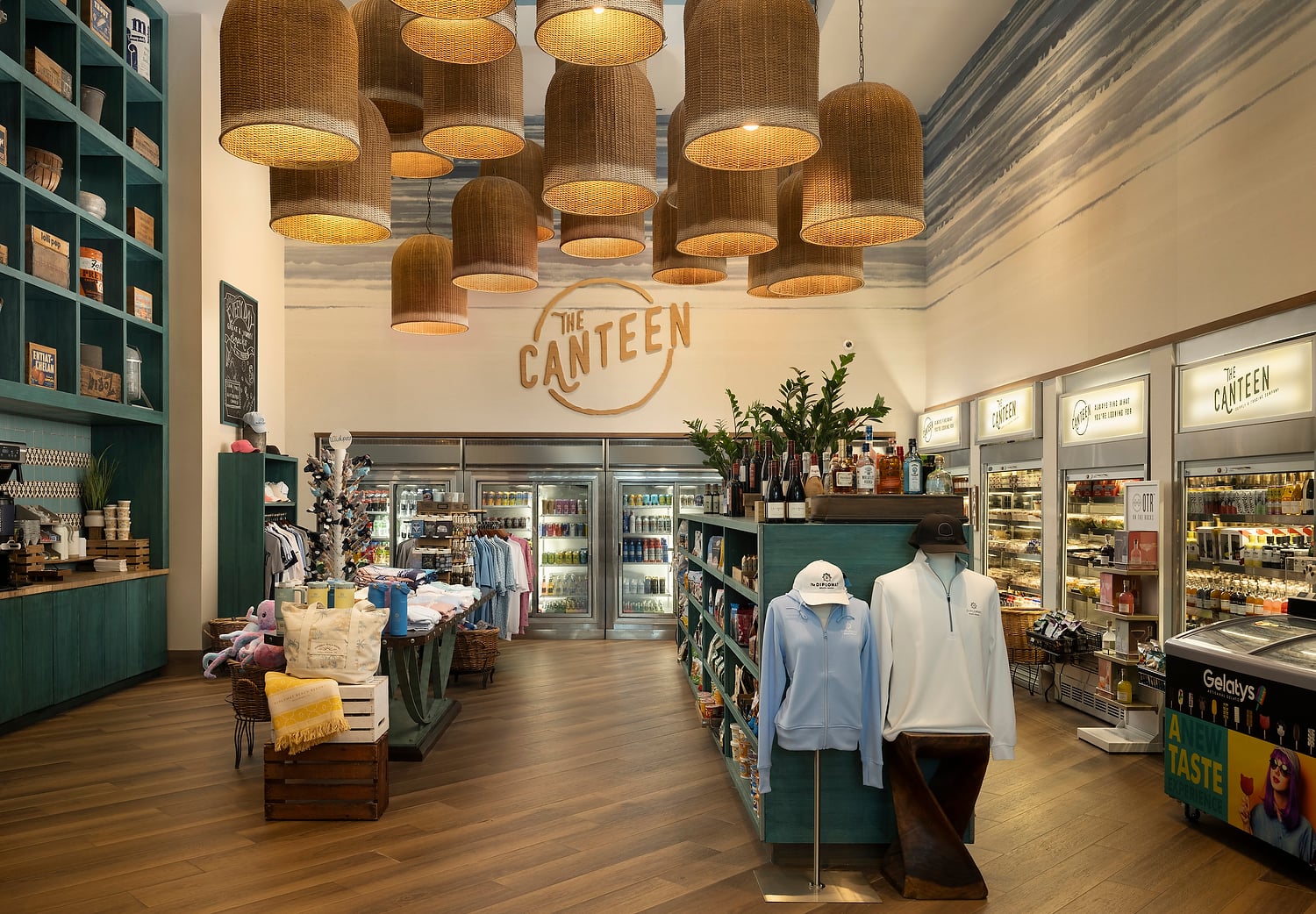 A bright convenience store interior displays apparel, beverages, and snacks. Wicker pendant lights illuminate teal shelves and wooden floors. Text found: THE CANTEEN, Gelatys, A NEW TASTE, ALWAYS FIND WHAT YOU'RE LOOKING FOR, OTR, ENTIRELY AMERICAN, THE DIPLOMAT, The Canteen.
