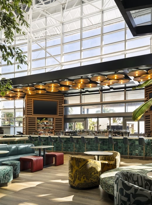 A modern, bright bar and lounge with a long green-tiled bar, diverse seating, and lush plants. It's under a large glass ceiling with ample natural light.
