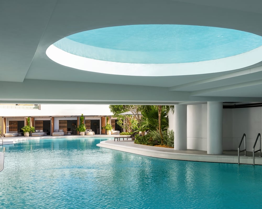 A large, bright blue swimming pool reflecting a circular ceiling opening. It's surrounded by lounge cabanas and tropical plants under a modern white architectural structure.