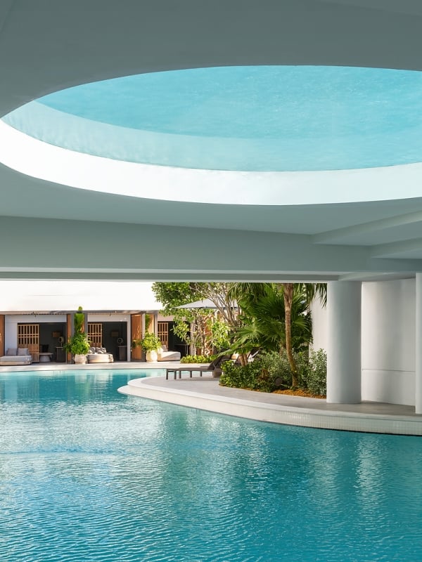 A large, bright blue swimming pool reflecting a circular ceiling opening. It's surrounded by lounge cabanas and tropical plants under a modern white architectural structure.