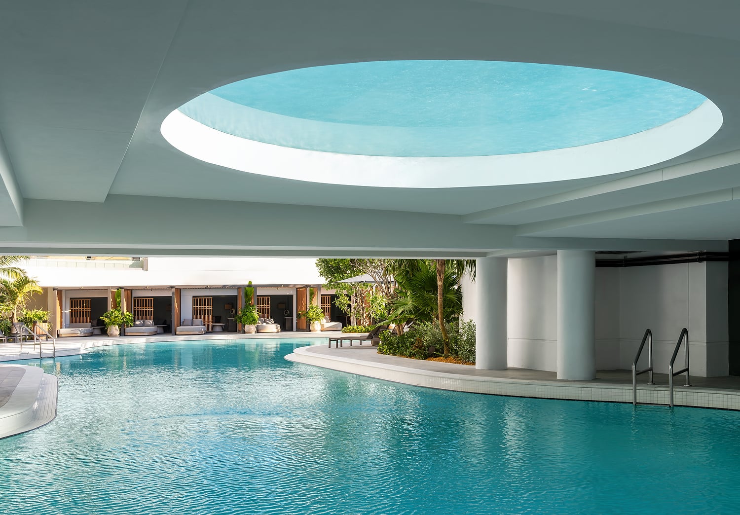 A large, bright blue swimming pool reflecting a circular ceiling opening. It's surrounded by lounge cabanas and tropical plants under a modern white architectural structure.
