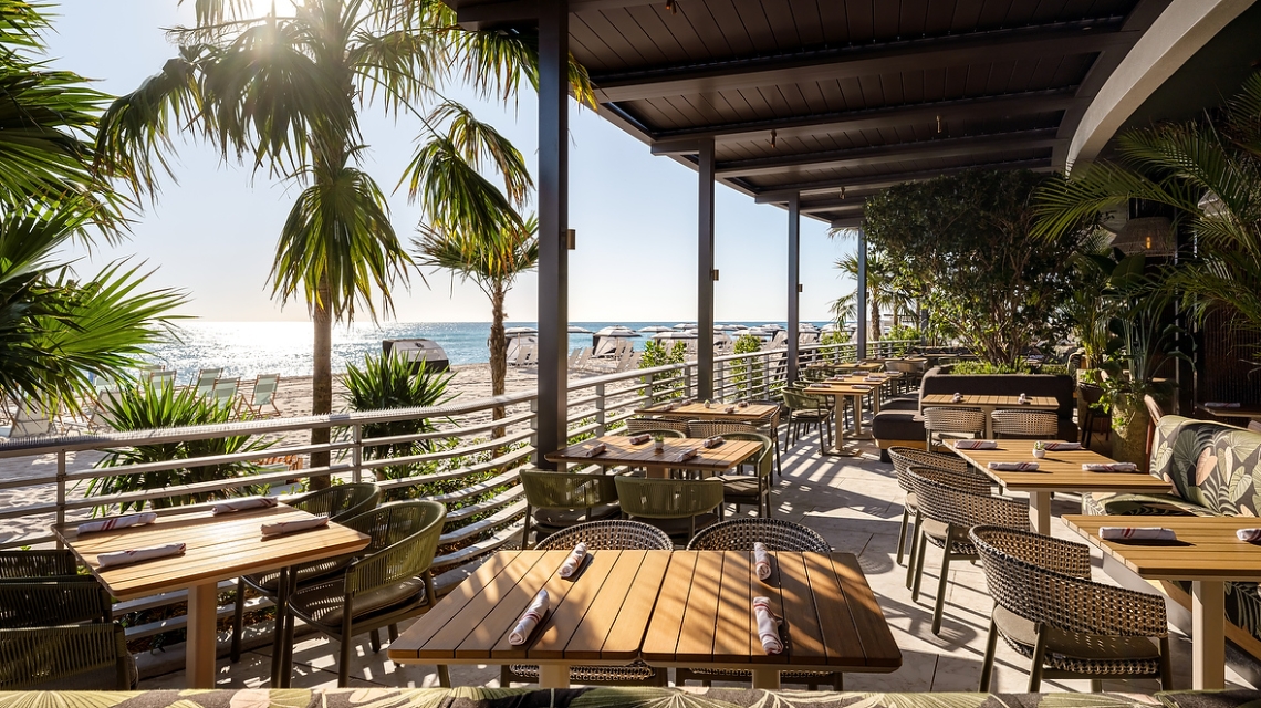 A shaded restaurant patio with multiple tables set for dining overlooks a sunny beach. Palm trees frame the ocean view with lounge chairs and cabanas.