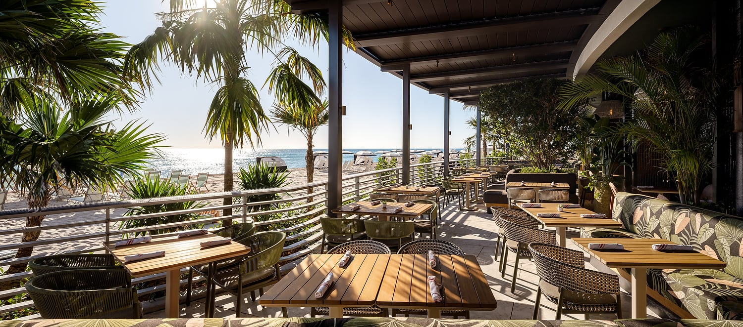 A shaded restaurant patio with multiple tables set for dining overlooks a sunny beach. Palm trees frame the ocean view with lounge chairs and cabanas.