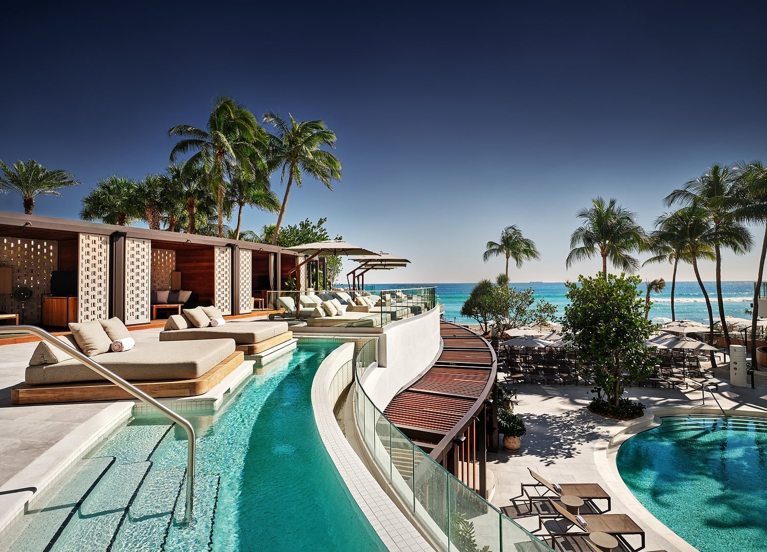 A luxury resort pool area features multiple pools, cabanas, and lounge chairs, overlooking a clear blue ocean with palm trees under a bright sky.