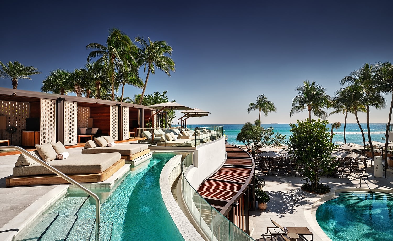 A luxury resort pool area features multiple pools, cabanas, and lounge chairs, overlooking a clear blue ocean with palm trees under a bright sky.