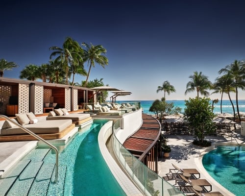 A luxury resort pool area features multiple pools, cabanas, and lounge chairs, overlooking a clear blue ocean with palm trees under a bright sky.