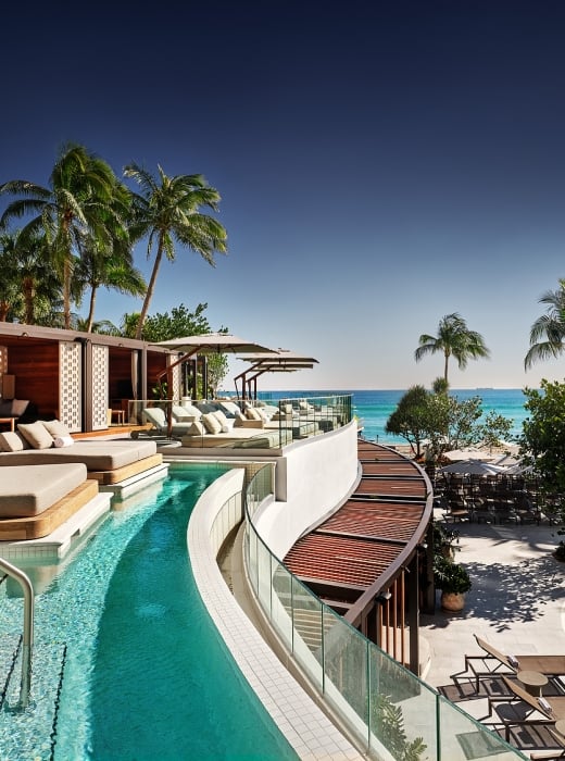 A luxury resort pool area features multiple pools, cabanas, and lounge chairs, overlooking a clear blue ocean with palm trees under a bright sky.