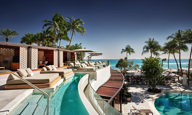 A luxury resort pool area features multiple pools, cabanas, and lounge chairs, overlooking a clear blue ocean with palm trees under a bright sky.
