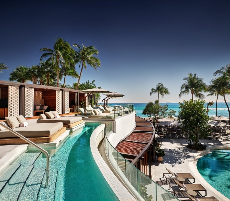 A luxury resort pool area features multiple pools, cabanas, and lounge chairs, overlooking a clear blue ocean with palm trees under a bright sky.