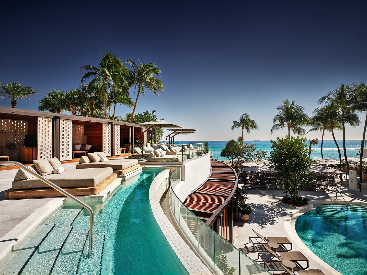 A luxury resort pool area features multiple pools, cabanas, and lounge chairs, overlooking a clear blue ocean with palm trees under a bright sky.