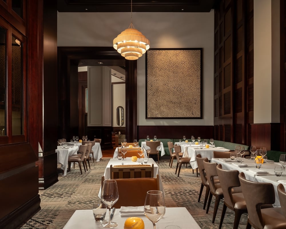 Set dining tables with white cloths and place settings fill an elegant, dimly lit restaurant. A large tiered chandelier illuminates dark wood paneling, a patterned rug, and wall art.