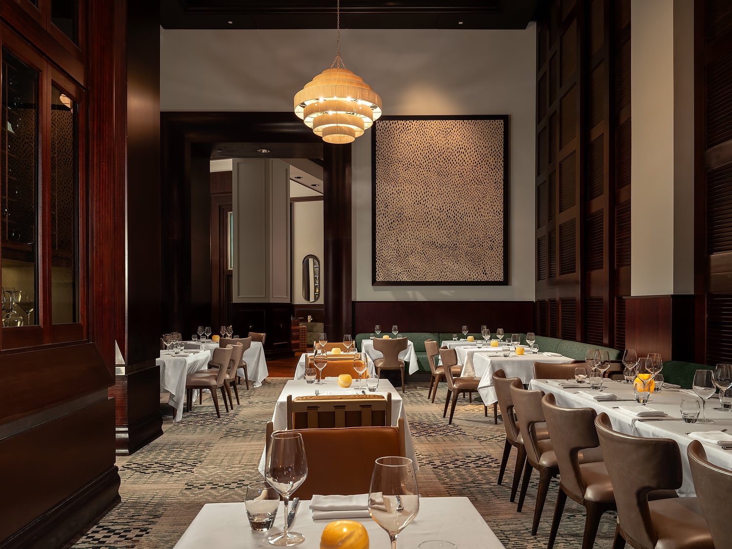 Set dining tables with white cloths and place settings fill an elegant, dimly lit restaurant. A large tiered chandelier illuminates dark wood paneling, a patterned rug, and wall art.