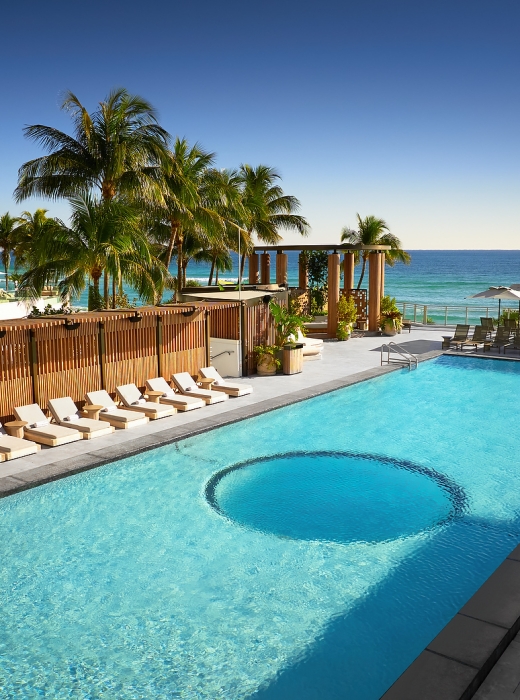 A long, light-blue swimming pool features a circular area, bordered by lounge chairs and tropical palm trees, overlooking the ocean under a clear sky.