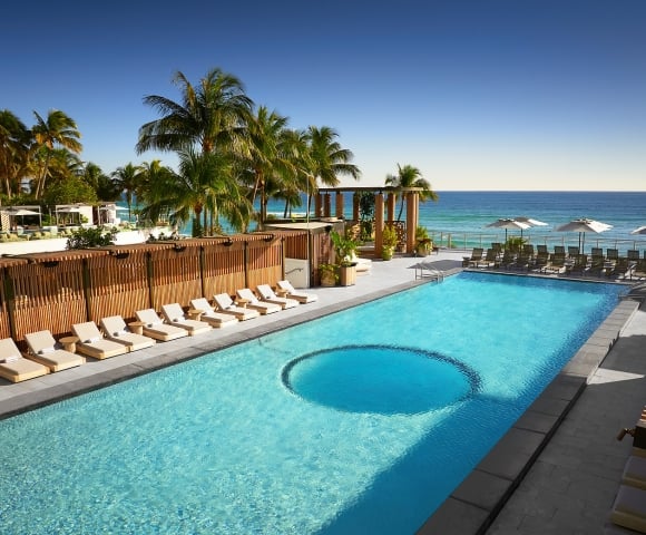 A long, light-blue swimming pool features a circular area, bordered by lounge chairs and tropical palm trees, overlooking the ocean under a clear sky.